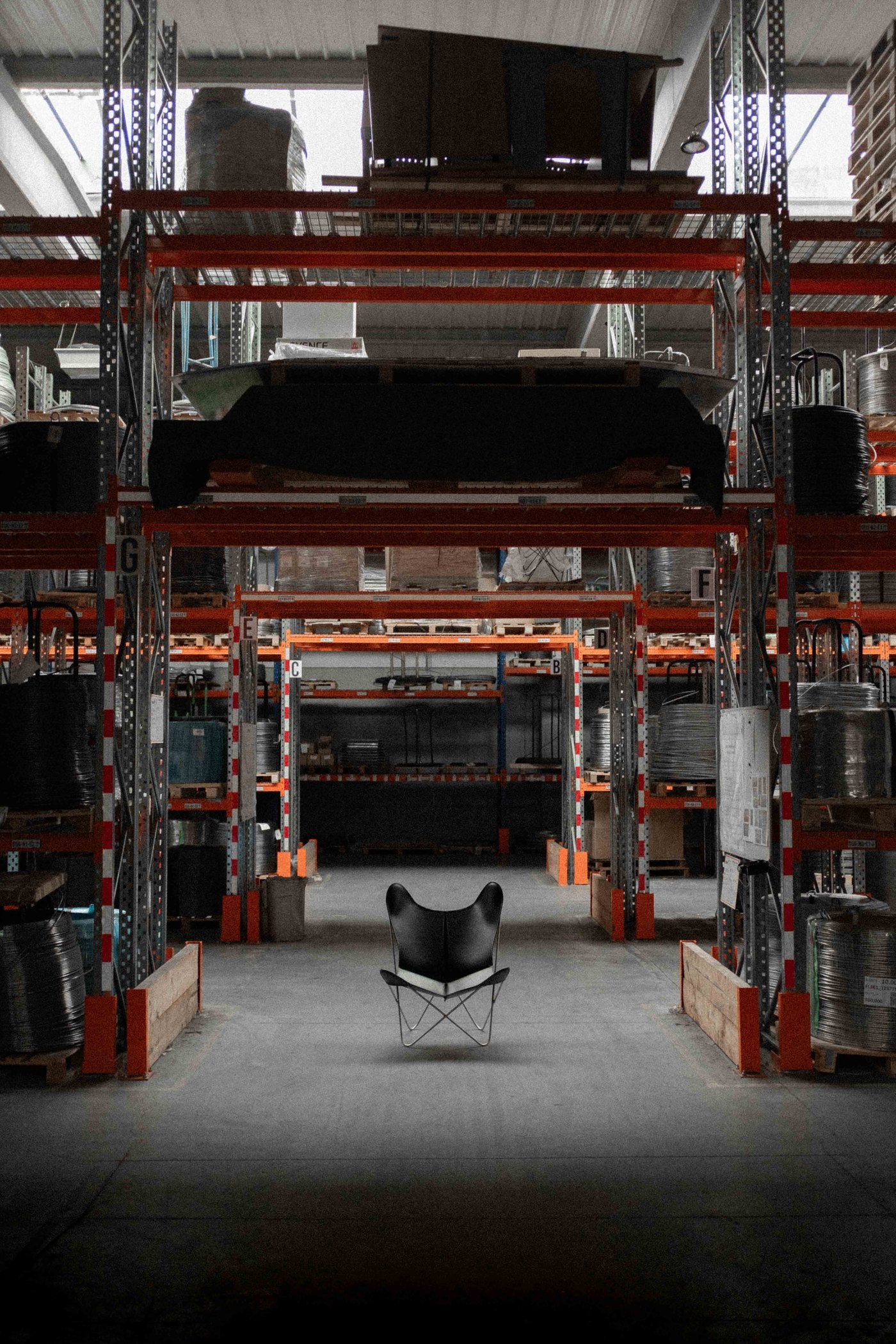 Fauteuil papillon AA Butterfly© en cuir noir posé seul dans l'allée centrale d'un entrepôt de manufacture, entre les racks d'acier